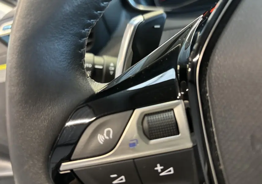 Gros plan sur les commandes noires et chrome du volant cuir de la Peugeot 208 blanche, côté gauche.