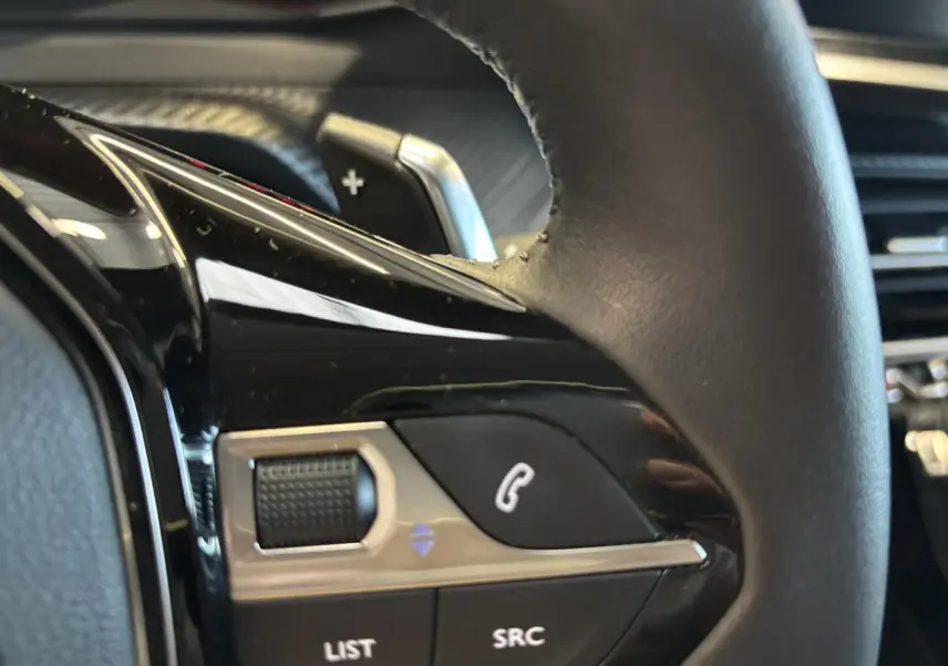 Gros plan sur commandes multimédia noires et chrome sur le volant cuir de la Peugeot 208 blanche, côté gauche.