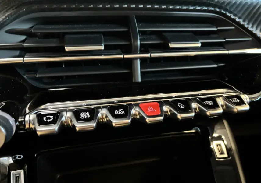 Gros plan sur la rangée de boutons chromés et noirs avec bouton rouge d’alerte, tableau de bord noir brillant Peugeot 208 blanc.