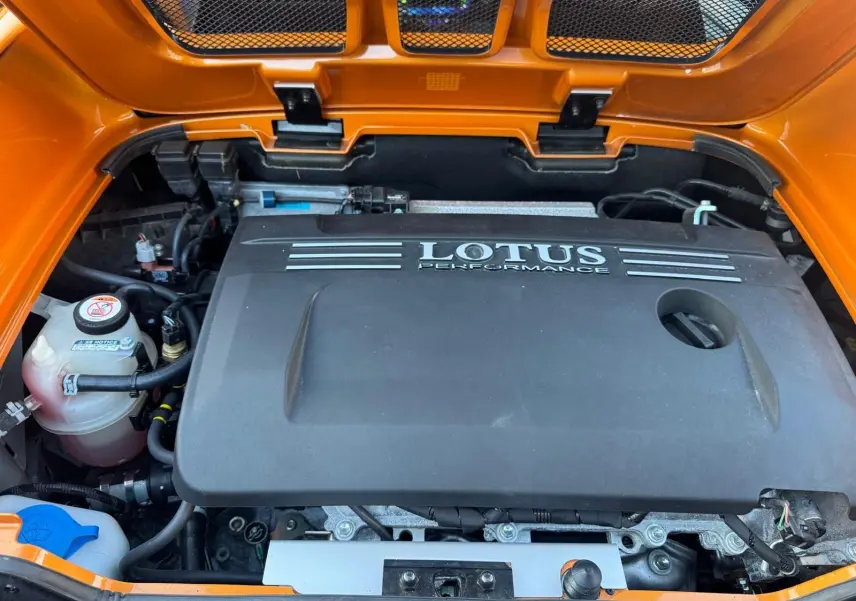 Vue rapprochée du compartiment moteur ouvert d'une Lotus Elise 3 Cup 250 orange, avec le cache moteur noir marqué "LOTUS PERFORMANCE".