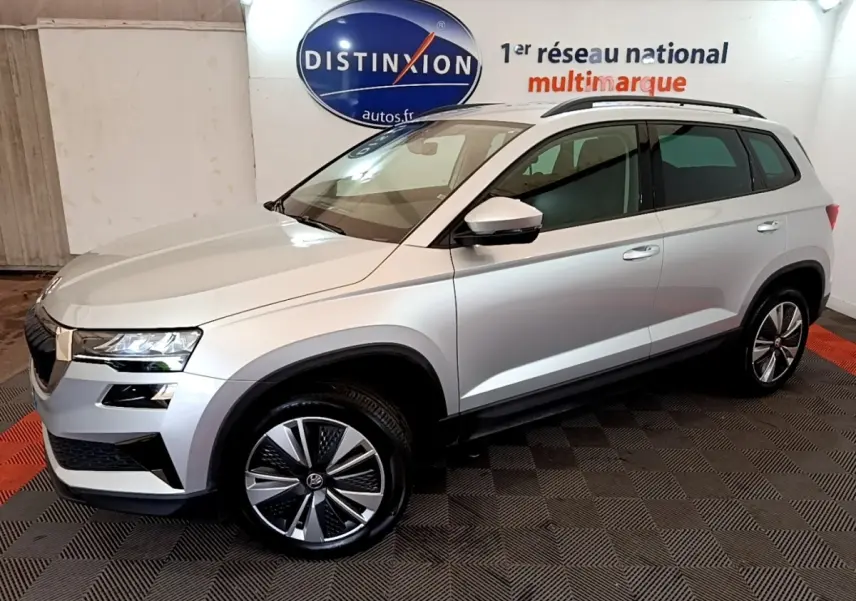 Vue 3/4 avant droite d’un Skoda Karoq gris clair 2022 avec jantes alliage et barres de toit noires.