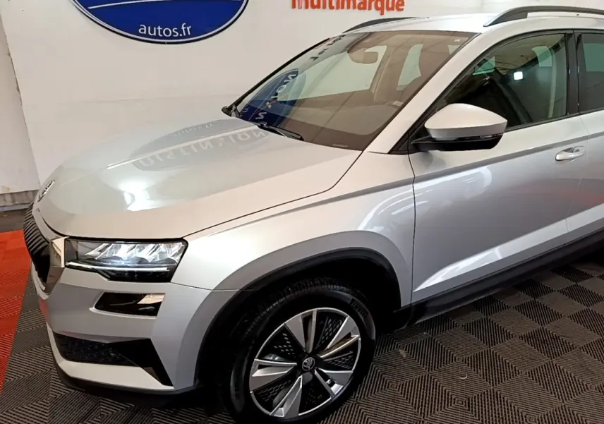 Vue 3/4 avant droit d'un Skoda Karoq gris clair avec jantes alliage et phares LED allumés en intérieur showroom.