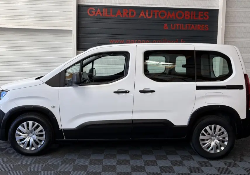 Profil droit d'un Peugeot Rifter blanc 2021 avec protections noires sur bas de caisse et jantes argentées, stationné en intérieur.