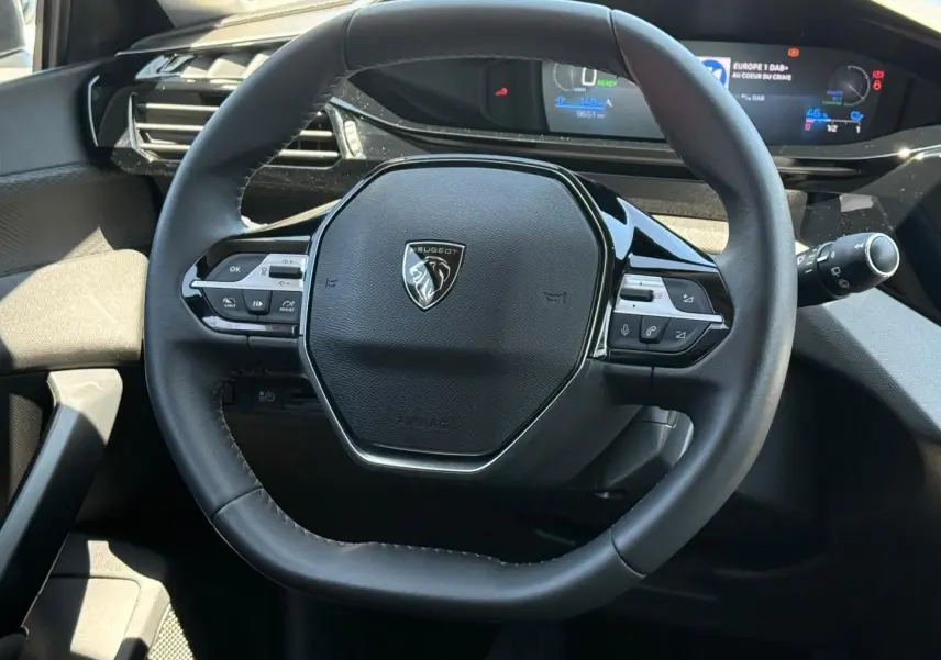 Vue rapprochée du volant noir multifonction de la Peugeot e-308 2025 avec tableau de bord digital en arrière-plan.
