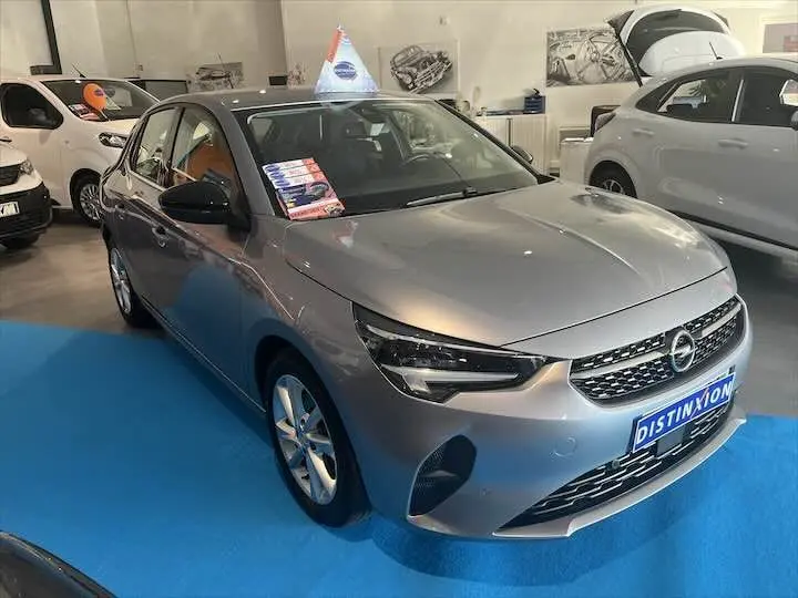 Opel Corsa gris métal vue 3/4 avant droit en showroom avec affiches sur le pare-brise et sol bleu.