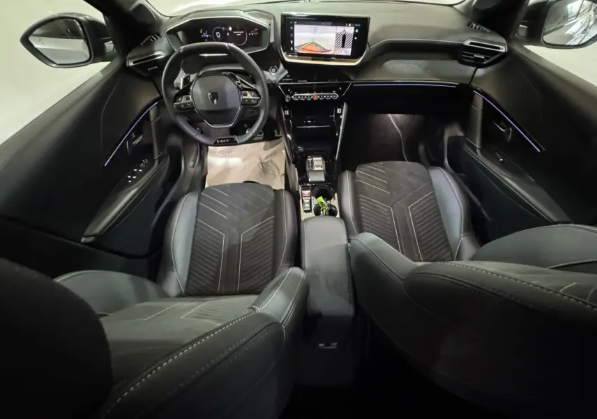 Vue intérieure avant de la Peugeot 208 Hybrid GT 2025, tableau de bord noir avec écran tactile et éclairage d'ambiance bleu.