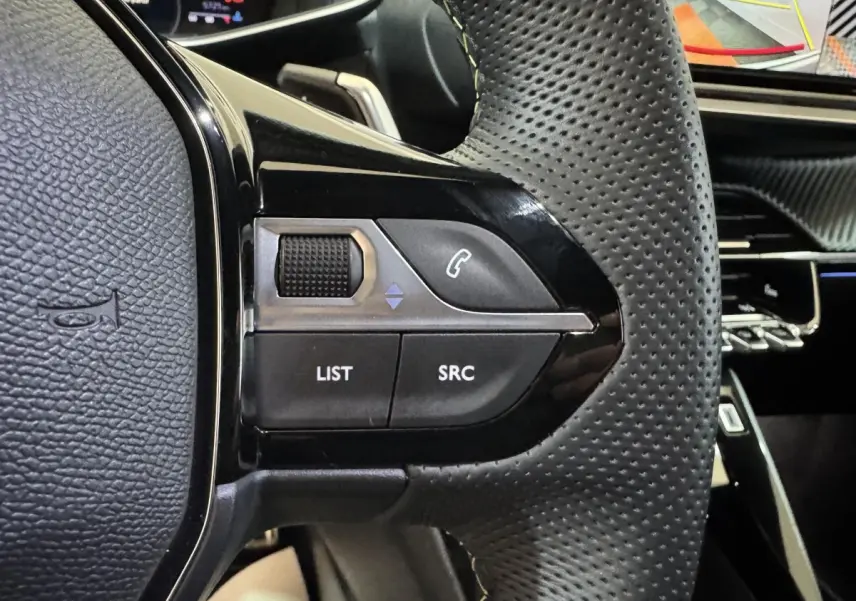 Gros plan sur les commandes gauche du volant en cuir perforé noir du Peugeot 208 Hybrid gris clair, avec tableau de bord en arrière-plan.