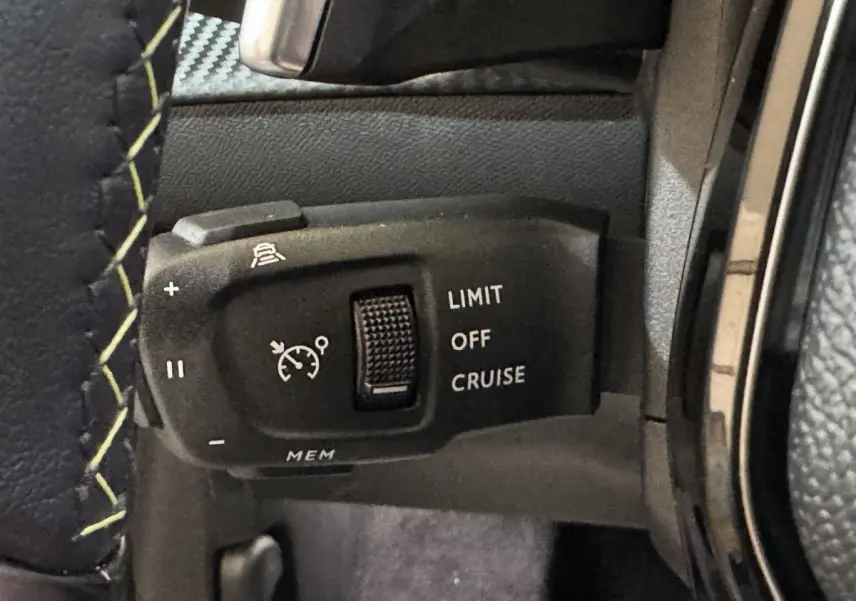 Gros plan sur la commande de régulateur de vitesse au volant cuir noir avec surpiqûres jaunes d’une Peugeot 208 Hybrid 136 eDCS6 GT gris clair.