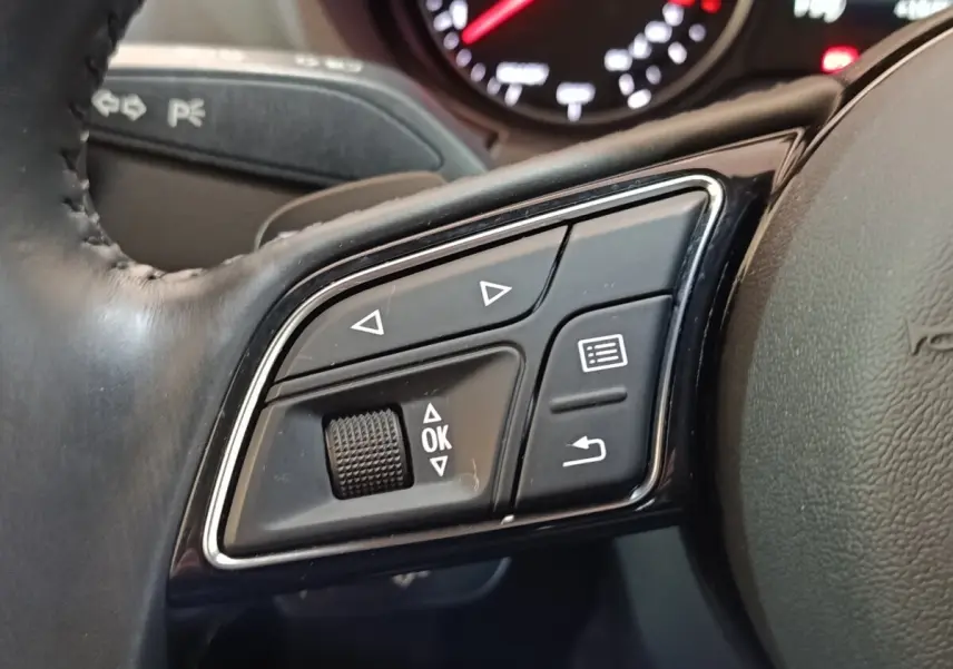 Gros plan sur les commandes noires multifonctions du volant cuir d'une Audi A3 blanche, avec tableau de bord en arrière-plan flou.