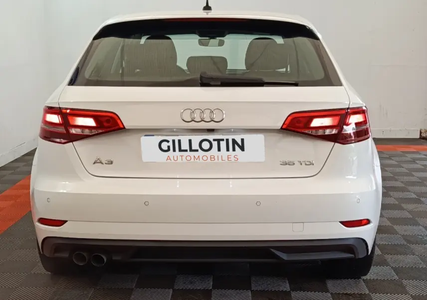 Vue arrière d'une Audi A3 35 TDI blanche avec feux allumés et double sortie d'échappement à gauche.