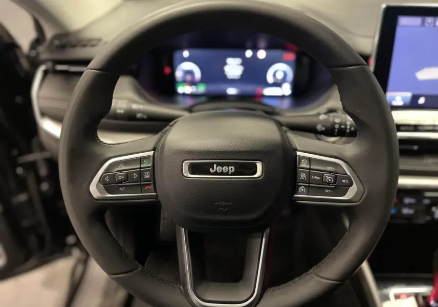 Vue rapprochée du volant noir du Jeep Compass 2024 avec commandes intégrées et tableau de bord numérique flou en arrière-plan.