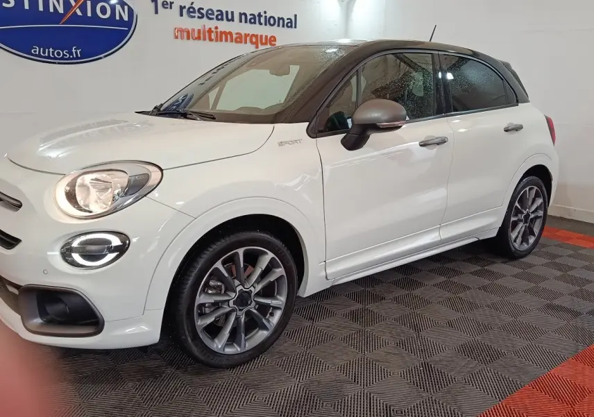 Vue 3/4 avant droit d'une FIAT 500X blanche avec toit noir et jantes alliage, en intérieur sur sol damier.