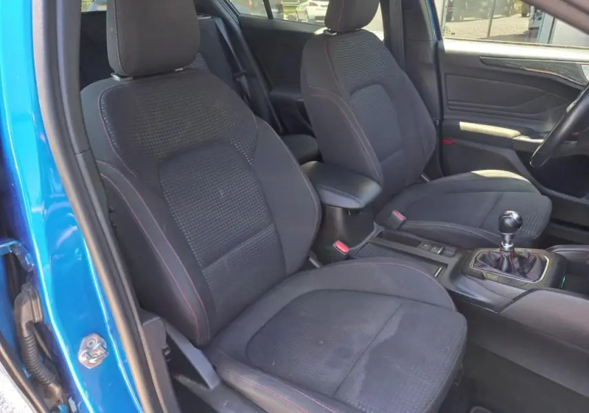 Vue intérieure côté conducteur d'une Ford Focus bleu 2019, sièges noirs avec surpiqûres rouges et levier de vitesse manuel.