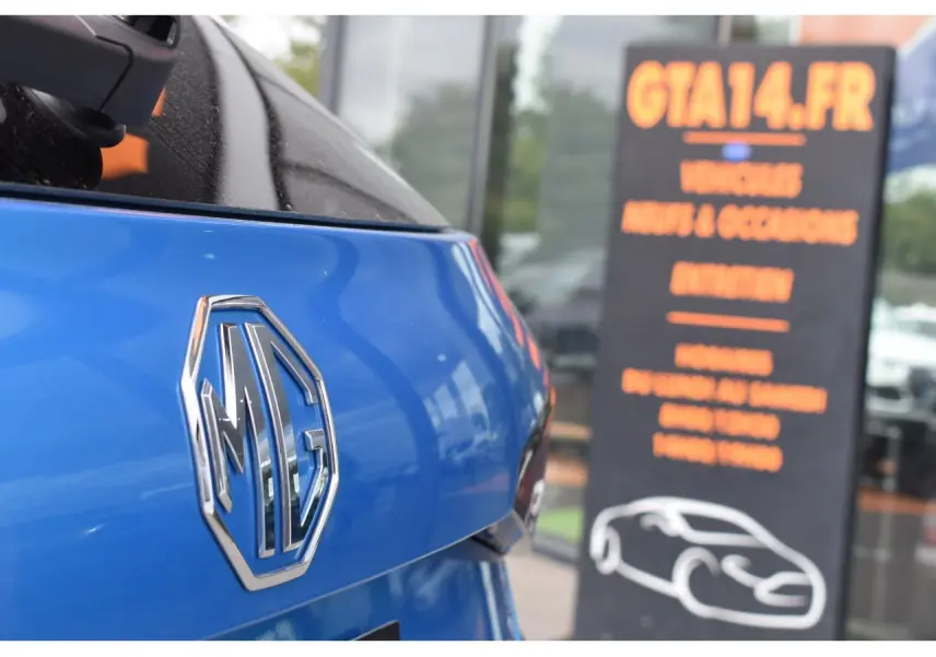 Gros plan sur le logo MG à l'arrière d'une MG3 1.5 HYBRID+ 195 Luxury bleu métallisé, avec reflet du ciel et panneau flou en arrière-plan.
