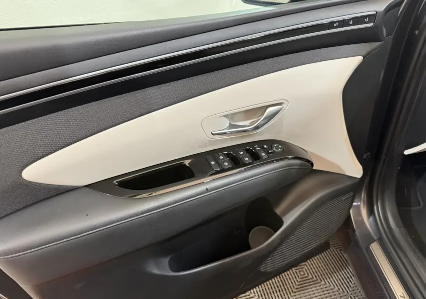 Vue intérieure du panneau de porte avant gauche du Hyundai Tucson gris foncé, avec commandes de vitres et réglages sièges.