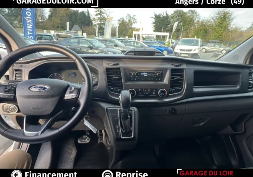 Vue intérieure avant du Ford Transit Custom blanc 2023, montrant le volant cuir multifonctions et la console centrale avec boîte auto.