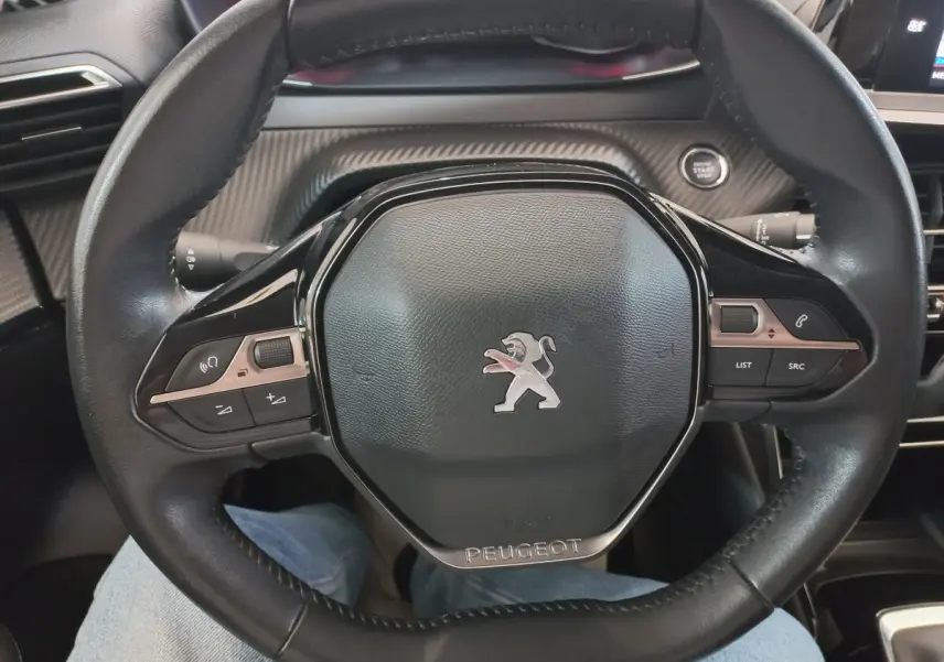 Vue rapprochée du volant cuir noir de la Peugeot 208 PureTech 100 S&S Allure avec commandes intégrées et bouton démarrage.