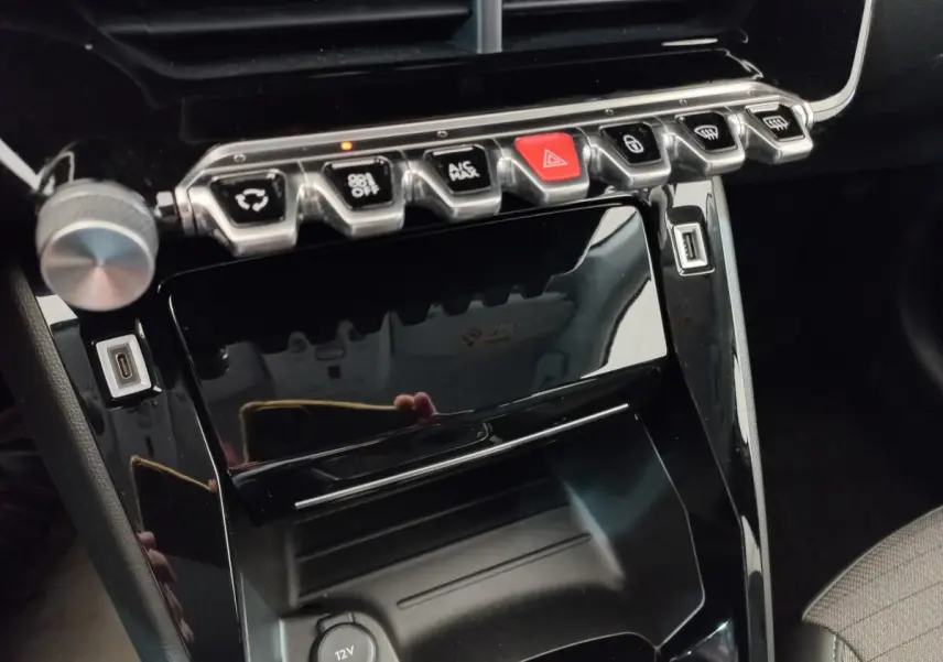 Gros plan sur la console centrale noire brillante de la Peugeot 208 PureTech 100 S&S Allure avec boutons de commande et ports USB.