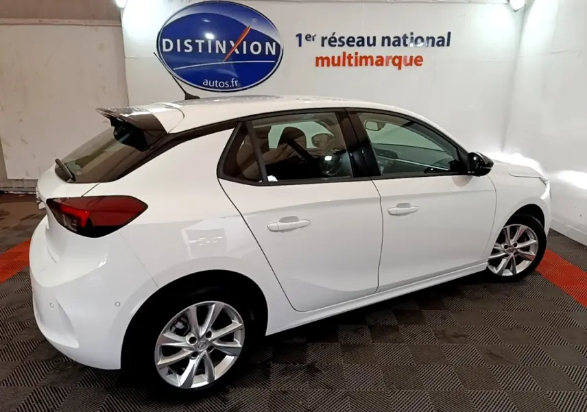 Vue 3/4 arrière droite d'une Opel Corsa blanche 2023 avec jantes alliage et spoiler de toit dans un showroom.