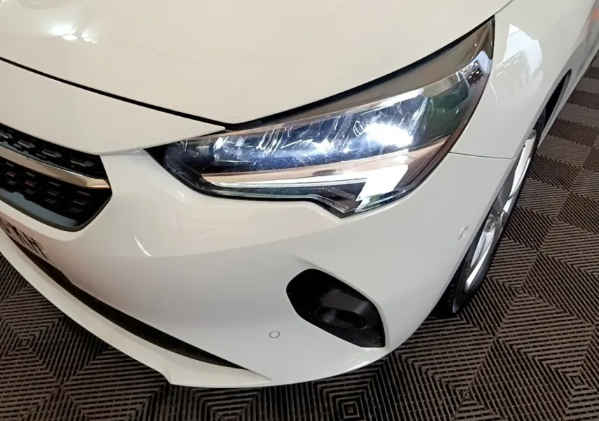Vue 3/4 avant droit d'une Opel Corsa blanche 2023, mettant en valeur le phare LED et le pare-chocs avec capteurs de stationnement.