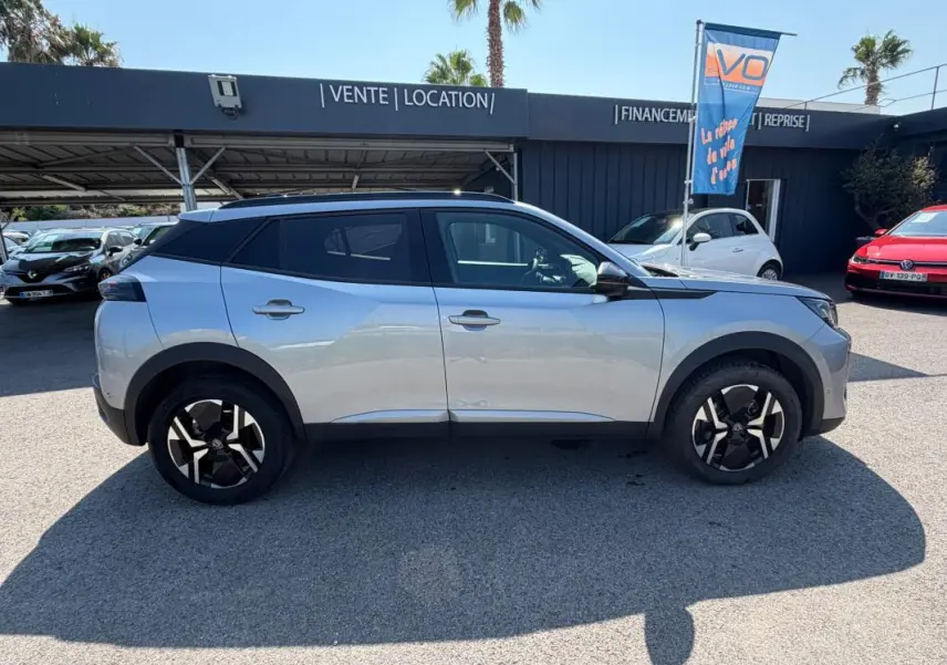 Profil côté gauche d’un Peugeot 2008 Hybrid 145 gris 2025 avec jantes noires et toit noir, stationné en extérieur.