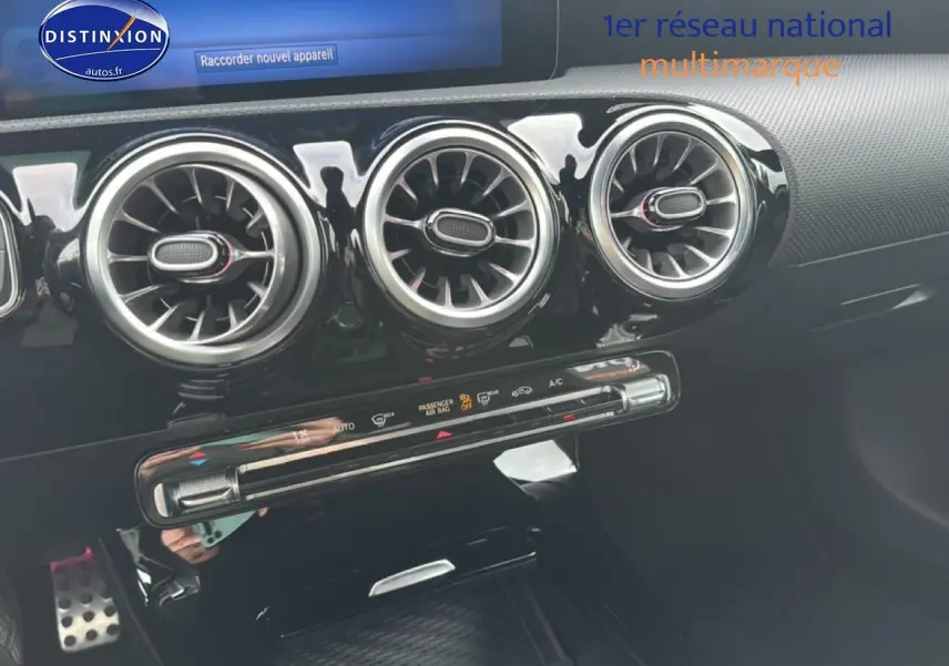 Gros plan sur la console centrale noire brillante avec trois aérateurs ronds et commandes climatisation d'une Mercedes Classe A bleue.