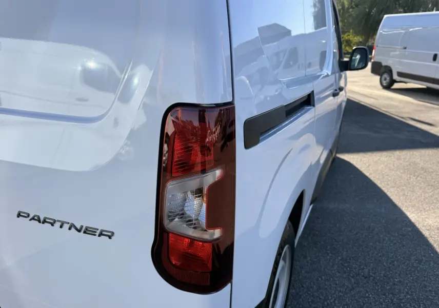 Vue arrière côté droit d’un Peugeot Partner blanc avec feu arrière et logo visible sur une route ensoleillée.