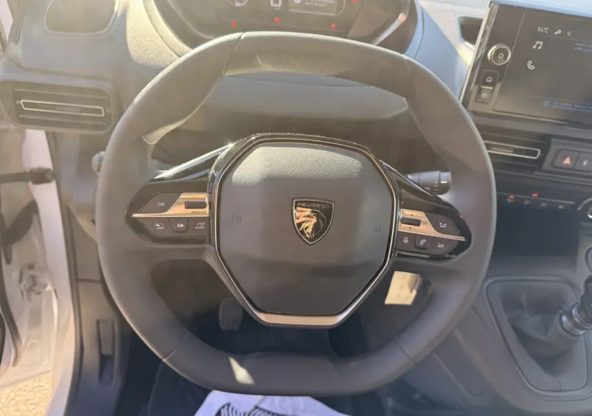 Vue rapprochée du volant multifonctions noir avec logo Peugeot au centre dans l'habitacle du Partner blanc 2026.
