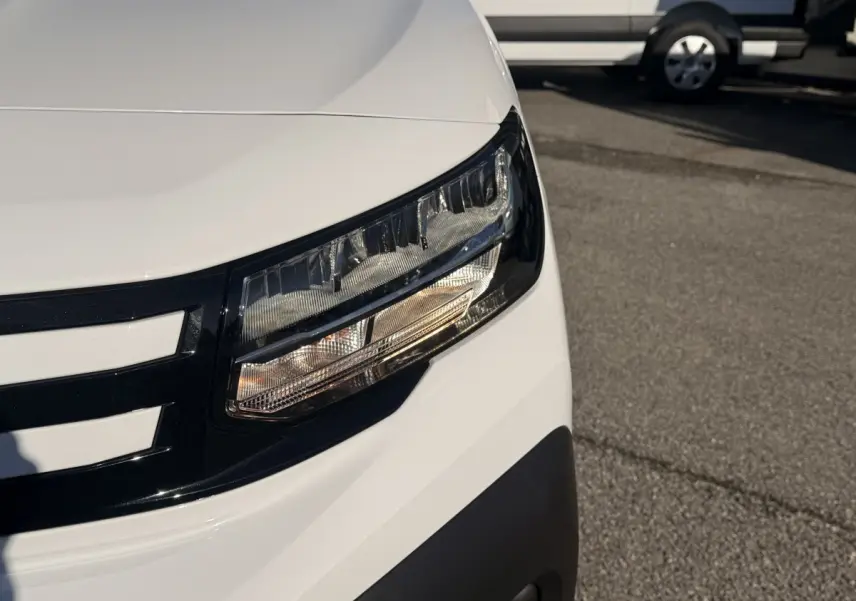 Gros plan sur le phare avant droit du Peugeot Partner blanc, soulignant la calandre noire et le pare-chocs gris.