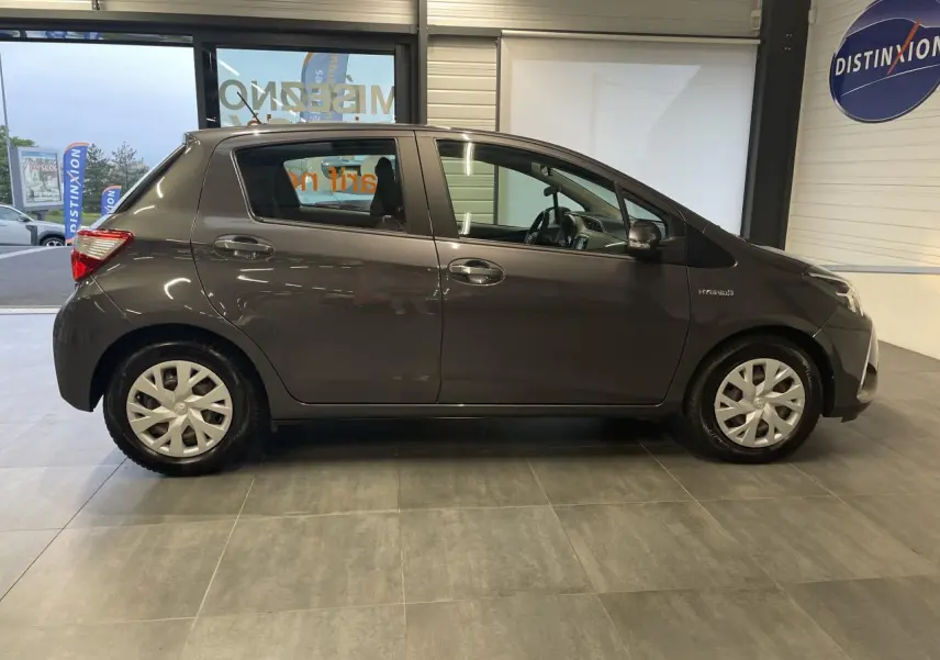 Profil côté gauche d'une Toyota Yaris hybride gris foncé métallisé, avec enjoliveurs 15 pouces et calandre noir laqué visible en showroom.