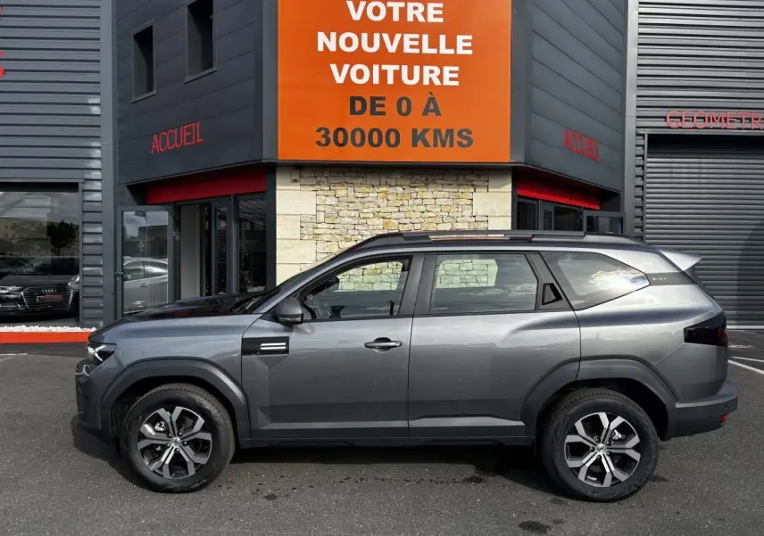 Profil côté gauche d'un Dacia Bigster gris foncé 2025 avec barres de toit noires, stationné devant un bâtiment commercial.
