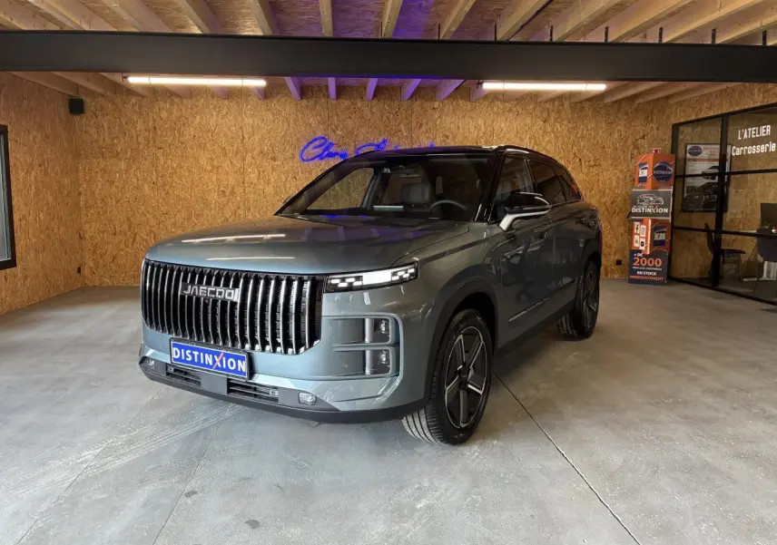 SUV JAECOO J7 gris olive en 3/4 avant droit, avec toit noir et calandre verticale imposante, en intérieur showroom.