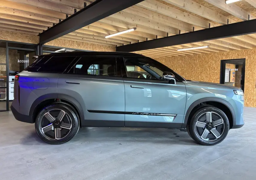 SUV JAECOO J7 gris olive avec toit noir, vu de profil côté gauche dans un garage en bois.