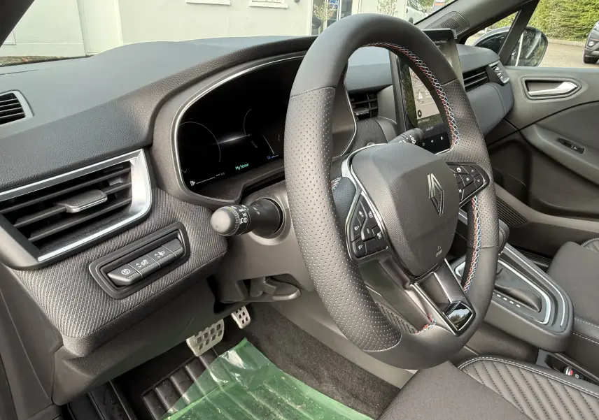 Vue intérieure côté conducteur de la Renault Clio V hybride 2025, volant cuir perforé avec surpiqûres tricolores et tableau de bord numérique.
