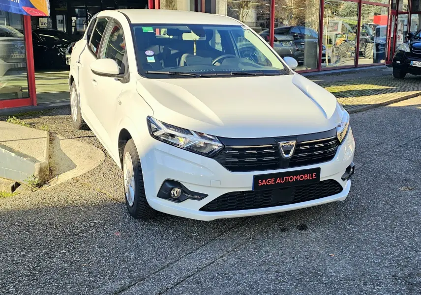 Vue 3/4 avant d'une Dacia Sandero III blanche glacier, avec calandre noire et phares LED, stationnée devant un showroom.