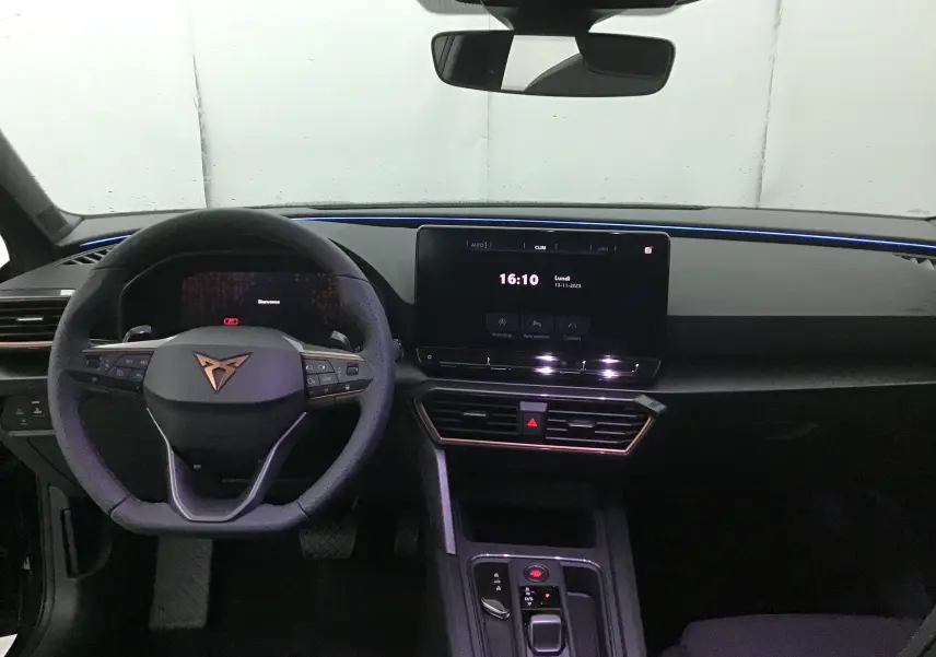 Vue intérieure du tableau de bord de la CUPRA Leon 2023 noire, avec volant multifonction, écran digital central et éclairage d'ambiance bleu.