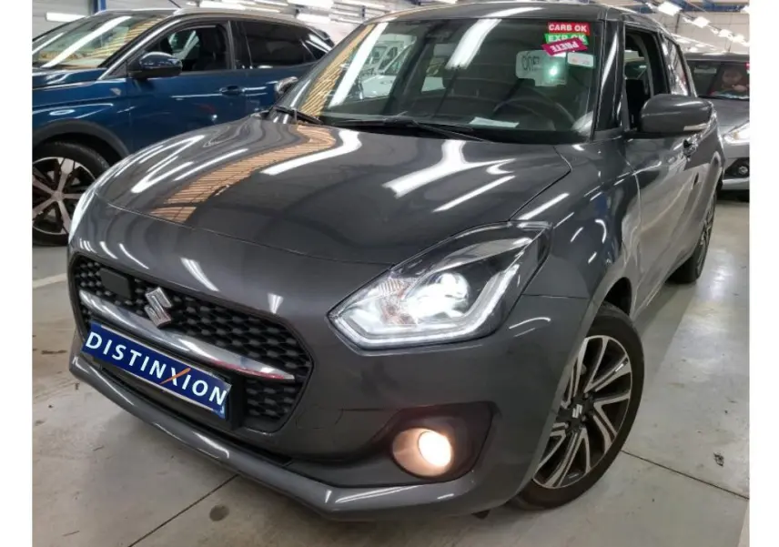 Suzuki Swift grise vue 3/4 avant droit, phares et antibrouillard allumés en intérieur showroom.