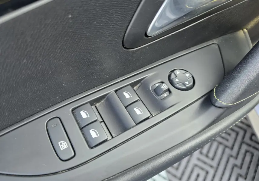 Gros plan sur les commandes de lève-vitres et rétroviseurs côté conducteur de la Peugeot 208 grise, avec surpiqûres vertes visibles.