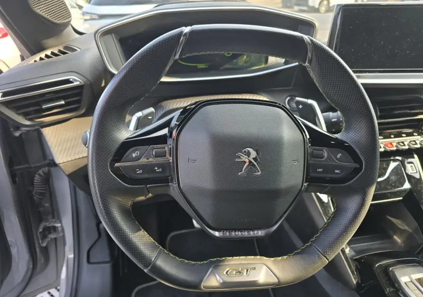 Vue rapprochée du volant cuir noir avec logo Peugeot et badge GT, intérieur gris et planche de bord carbone.