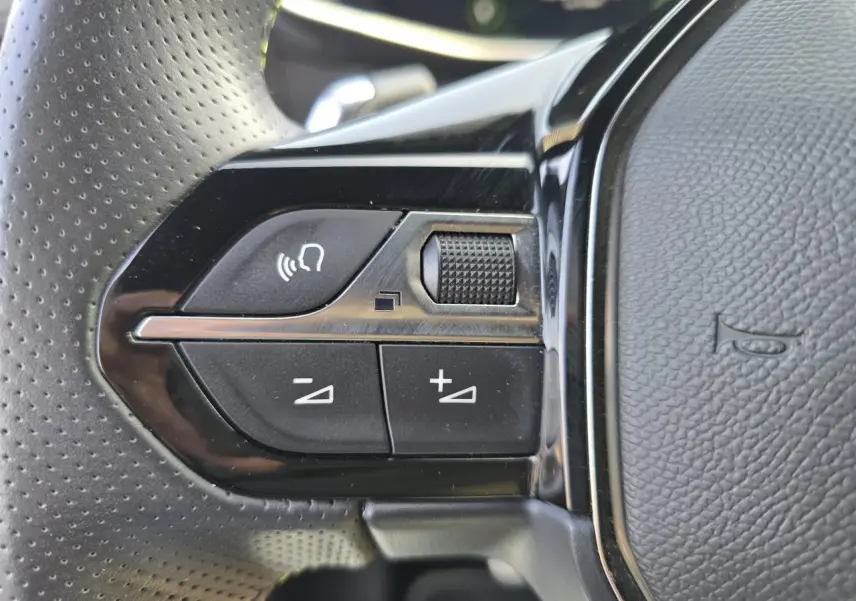 Gros plan sur les commandes de volume et reconnaissance vocale sur le volant noir perforé d'une Peugeot 208 grise 2023.