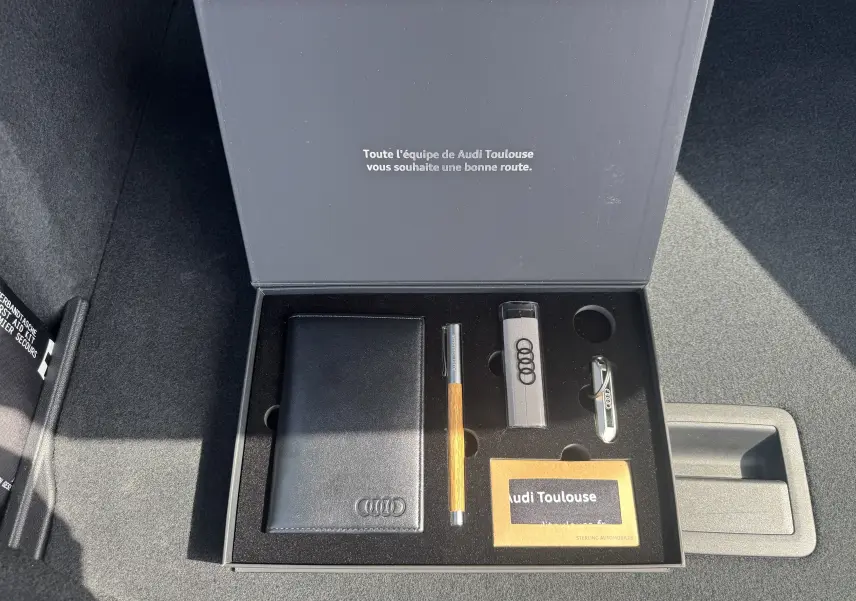 Boîte ouverte dans le coffre d’une Audi A3 Sportback gris, contenant porte-documents, stylo et clés Audi.