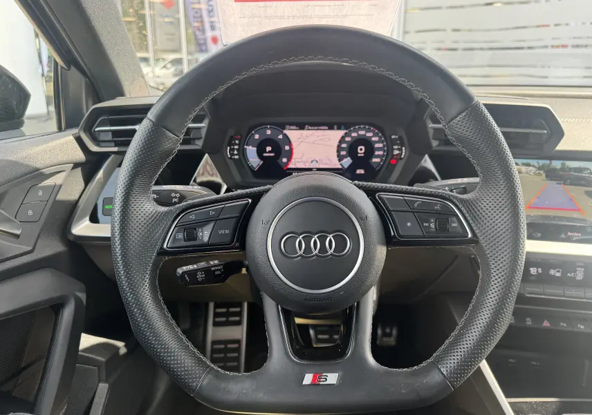 Vue centrée sur le volant noir perforé de l'Audi A3 Sportback TDI 2.0 S-Line 2023 avec tableau de bord numérique et caméra de recul.