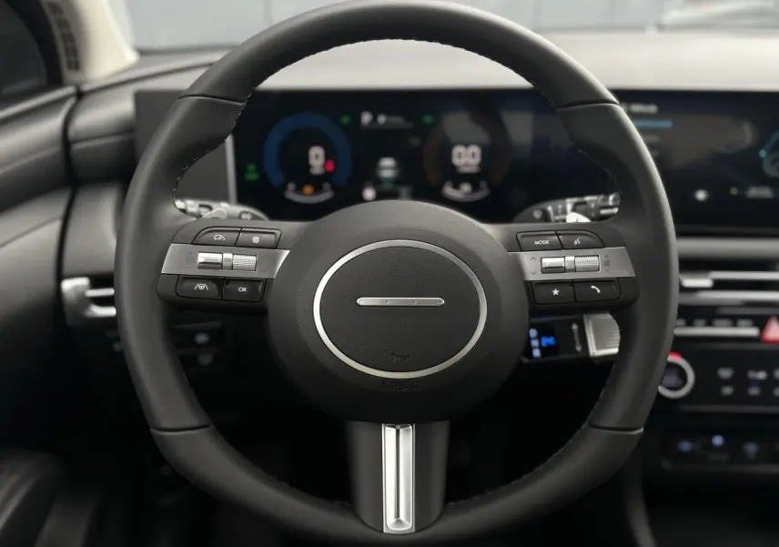 Vue rapprochée du volant noir du Hyundai Tucson 2026 avec commandes intégrées et tableau de bord numérique flou en arrière-plan.