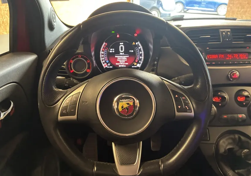 Vue intérieure centrée sur le volant cuir d'une Abarth 500 rouge de 2016, avec tableau de bord numérique et commandes au volant visibles.