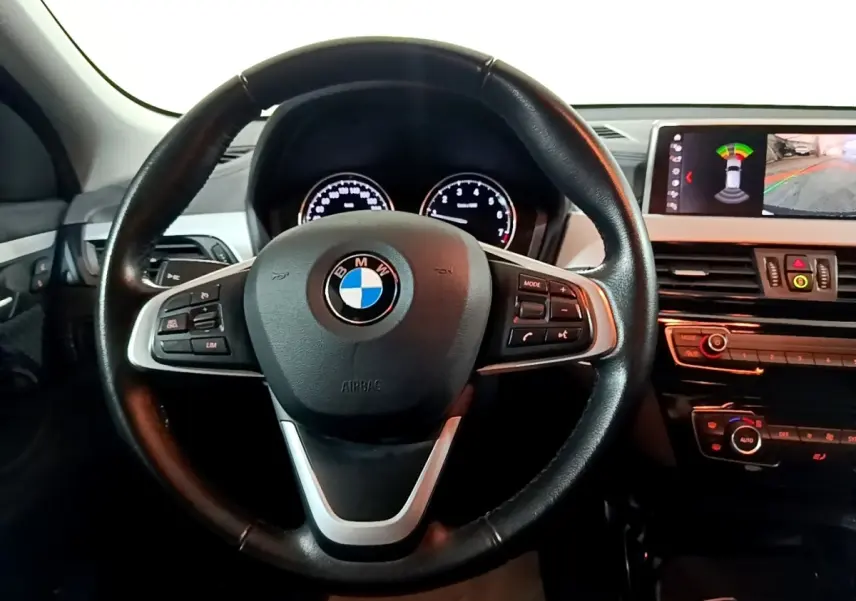 Vue intérieure centrée sur le volant cuir noir de la BMW X2 avec tableau de bord et écran d'aide au stationnement.