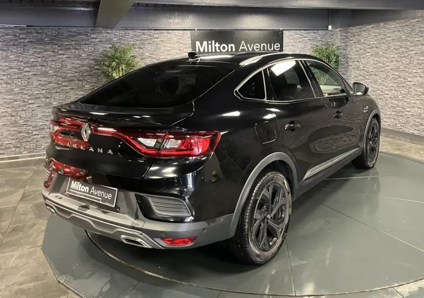 Vue 3/4 arrière droite d'un Renault Arkana noir brillant avec jantes noires et feux arrière LED allumés en intérieur showroom.