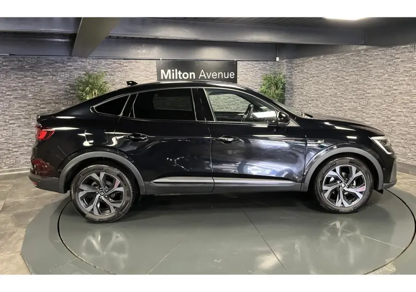 Vue de profil côté gauche d'un Renault Arkana noir métallisé avec jantes alliage et toit plongeant en intérieur showroom.