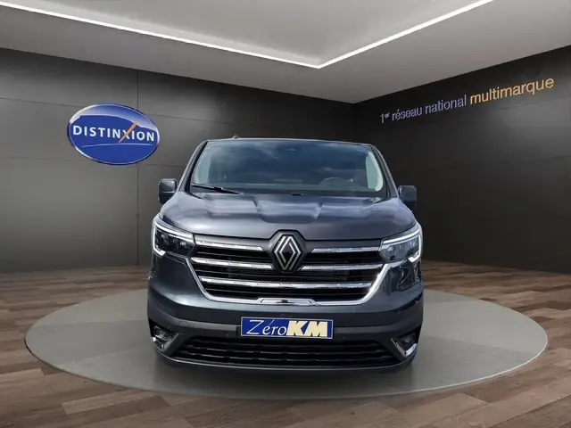 Vue de face d’un Renault Trafic Fourgon gris foncé, avec calandre chromée et phares LED allumés, en showroom.