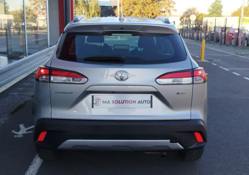 Vue arrière d'une Toyota Corolla Cross gris minéral, avec feux allumés et logo hybride visible sur un parking urbain.