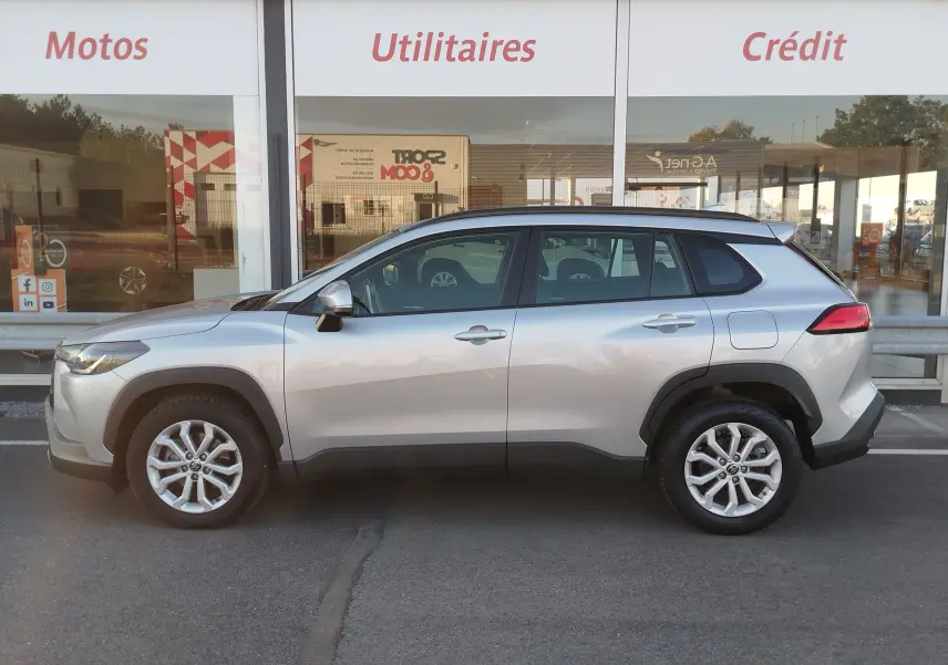 Vue de profil côté gauche d'un Toyota Corolla Cross gris minéral, SUV hybride avec jantes alliage et protections noires.